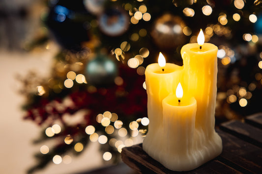 Three lit cream-colored pillar candles with a textured, ribbed design, set on a dark wooden surface with a blurred Christmas tree and warm bokeh lights in the background.