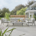 Outdoor patio set with sofa, chairs, and coffee table on a deck with a modern house and garden in the background.