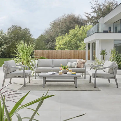 Outdoor patio set with sofa, chairs, and coffee table on a deck with a modern house and garden in the background.