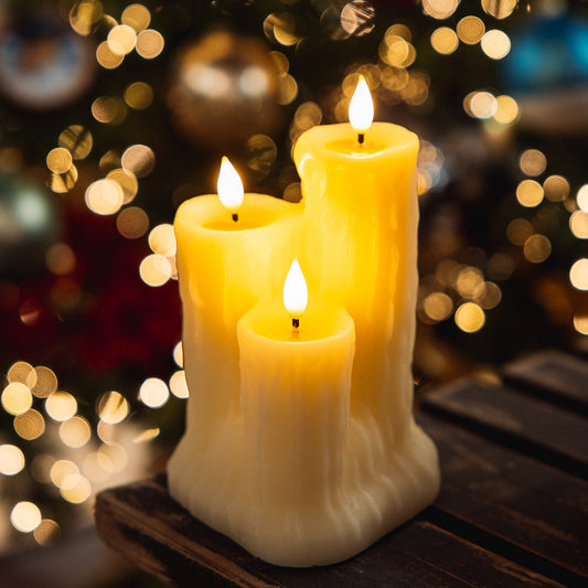 Three lit cream-colored pillar candles with a textured, melting-wax design, displayed on a dark wooden surface with a festive, out-of-focus Christmas tree and bokeh lights in the background.